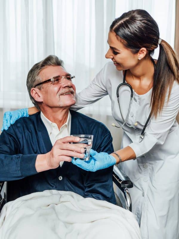 An older man in a wheelchair smiles at the nurse-assistant, she hands him a glass of water. Nursing home, help for the elderly.